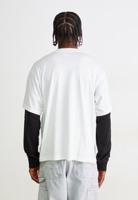 Tricou alb din bumbac cu mânecă scurtă, suprapus peste un tricou negru cu mânecă lungă, având o croială lejeră și detalii de cusătură vizibile.