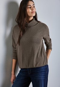 Brun, ribbad polotröja med en avslappnad passform och långa ärmar, i kombination med mörkblå jeans, stående mot en ljusgrå bakgrund.