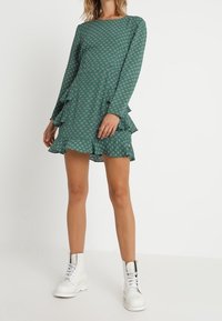 Vihreä kuvioitu mekko, jossa on pitkät hihat, röyhelöhelma ja pyöreä kaula-aukko, yhdistettynä valkoisiin nilkkabootseihin. Pehmeä kangas, rento muotoilu.