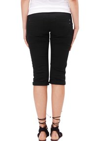 Pantalones capri negros de tela elástica, con dobladillos enrollados y bolsillos traseros, llevados con sandalias de tiras, fotografiados desde atrás.