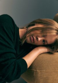 Modèle dans un pull noir reposant sur un coussin texturé beige, avec de longs cheveux partiellement couvrant son visage, montrant des traits doux et une pose détendue.