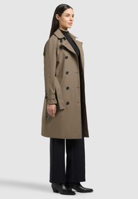 Donna con capelli lunghi e scuri che indossa un trench doppiopetto color cachi sopra un dolcevita nero, pantaloni neri a gamba larga e stivaletti neri.