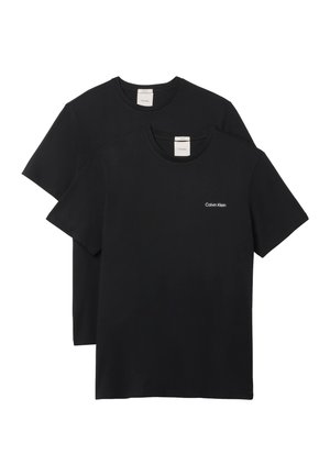Uomo in piedi che indossa una maglietta nera Calvin Klein, pantaloni neri, cintura nera e scarpe nere su uno sfondo grigio uniforme.