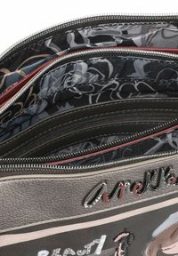 Schwarze synthetische Handtasche mit mehreren Reißverschlussfächern, strukturiertem grauem Außenmaterial und floral gemustertem Innenfutter, mit einem dekorativen Logo auf der Vorderseite.