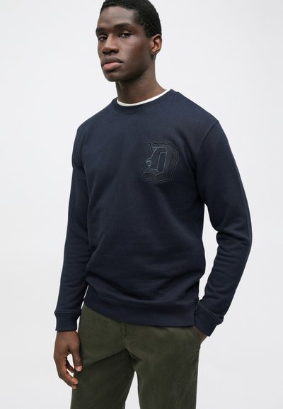 Mørkeblå sweatshirt med et tekstureret "D" logo på brystet, rund halsudskæring og ribbede manchetter og kant. Båret sammen med olivengrønne bukser.