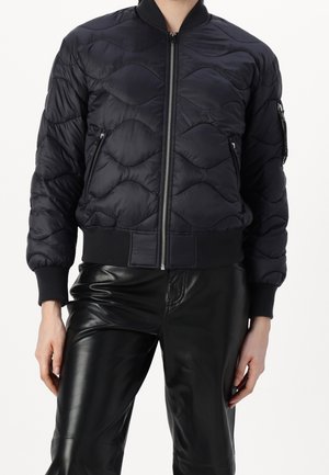 Personne portant une veste bomber noire matelassée avec des poches zippées et un pantalon en cuir noir, debout devant un fond uni.