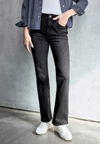 Jeans neri in denim a gamba svasata con vita media, dotati di cinque tasche e un leggero effetto slavato, abbinati a sneakers bianche su uno sfondo di cemento grigio.