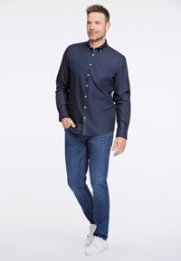 Camicia button-up blu navy con taschino sul petto, abbinata a jeans blu medio e sneakers bianche. Tessuto liscio, vestibilità sagomata.