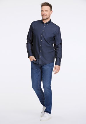 Marineblaue Button-Up-Bluse mit einer Brusttasche, kombiniert mit mittelblauen Jeans und weißen Sneakers. Glattes Material, taillierte Passform.