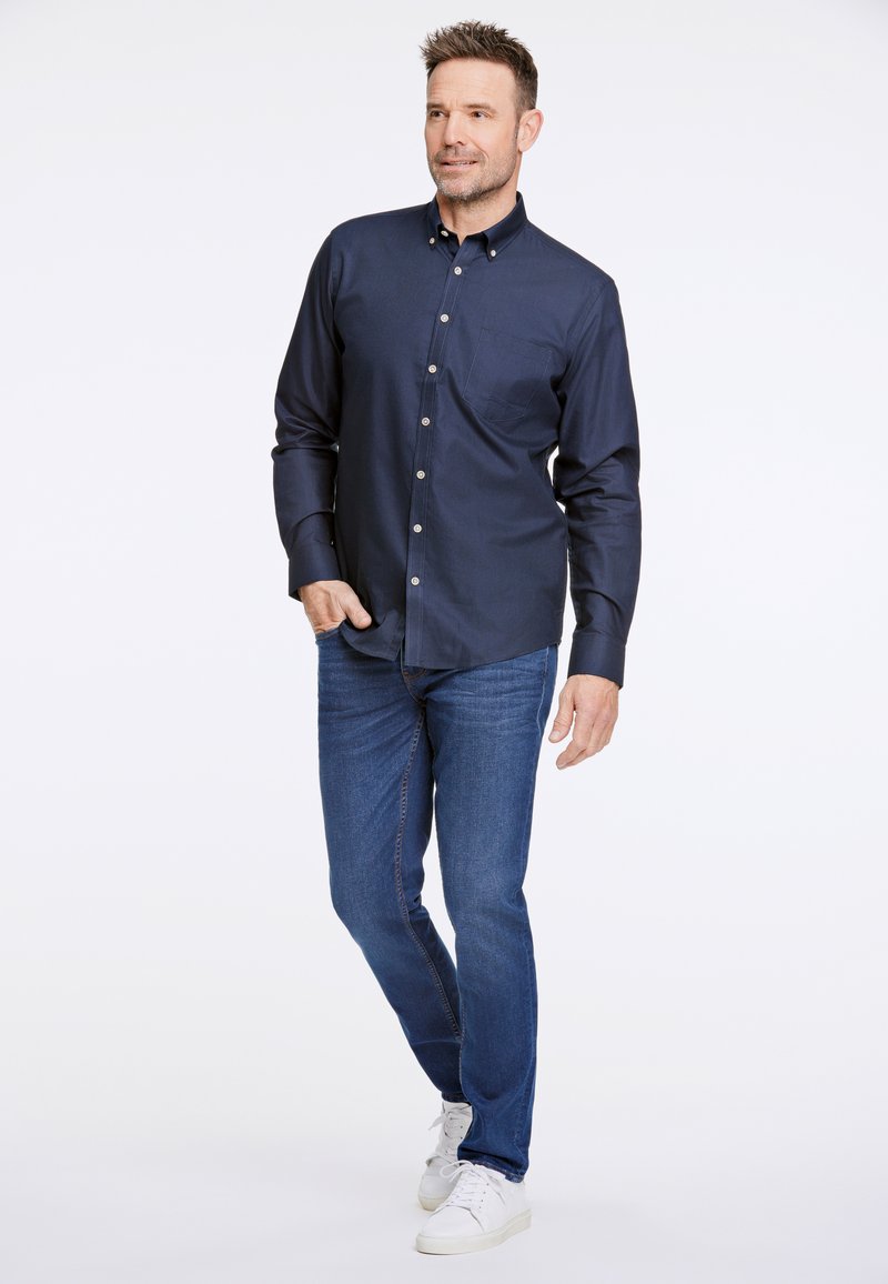 Camicia button-up blu navy con taschino sul petto, abbinata a jeans blu medio e sneakers bianche. Tessuto liscio, vestibilità sagomata.