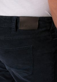 Mörkblå manchestertygbyxor med en läderpatch, bakficka med sömnadsdetaljer och en texturerad yta.