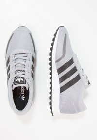 Grå träningsskor med ett nätöverdrag, svarta ränder och en vit sula. Har snören och en texturerad sula för grepp.