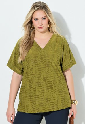 Lyshåret kvinde iført guldbøjleøreringe og en tekstureret olivengrøn V-hals bluse med korte ærmer, kombineret med mørke bukser.