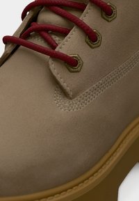 Εγγύς πλάνο μπεζ καστόρινου μποτακιού με κόκκινες κορδόνια διασχίζοντας ορειχάλκινα παπούτσια και σόλα από καουτσούκ γκαμ.