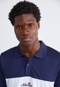 Marineblå polo t-skjorte med hvit aksent på brystet, med krage og knappdetaljering, laget av glatt stoff. Merkevaren logo er tydelig vist.