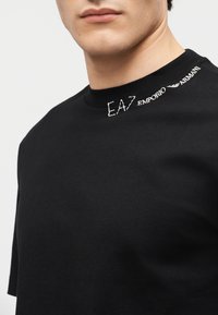 Κοντινό πλάνο ατόμου που φοράει μαύρο πουκάμισο με λευκό κείμενο "EA7 Emporio Armani" κεντημένο κοντά στον γιακά στη δεξιά πλευρά.