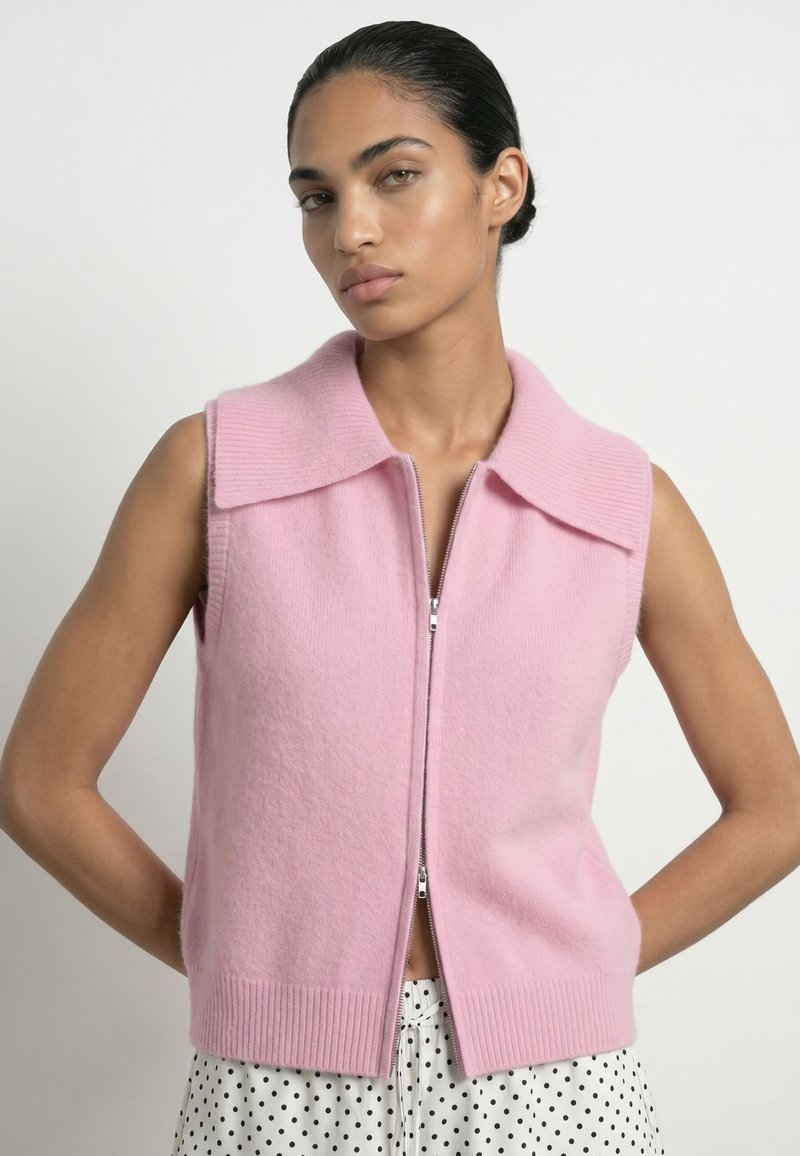 Femme portant un gilet sans manches rose zippé avec un large col sur une jupe blanche à pois noirs, posant avec les mains dans le dos.