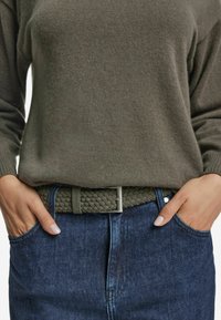 Flätad grön textilbälte med en spänne i silvermetal, bärs över en grå tröja och mörkblå denimjeans. Texturerat vävdetalj synlig.