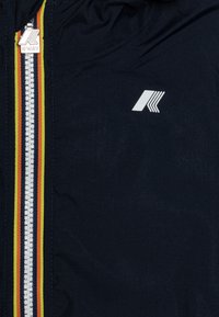 Veste K-Way navy avec une fermeture éclair blanche ornée de rayures rouge, jaune et verte, et un logo blanc sur la poitrine. Texture lisse, design léger.