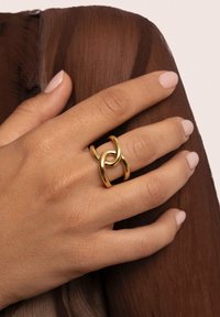 Goldener Metallring mit verflochtenen Schlaufen und einer polierten Oberfläche. Die Hand präsentiert einen hellbraunen Stoffhintergrund mit glatter Textur.
