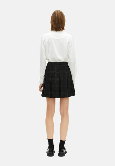 Witte blouse met lange mouwen en een rugpas, gecombineerd met een zwarte geruite geplooide rok. Zwarte schoenen en sokken maken de look compleet.