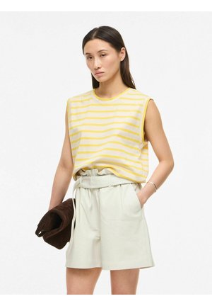 Femme portant un haut sans manches rayé jaune et blanc et un short beige clair taille haute, tenant une veste marron pliée.