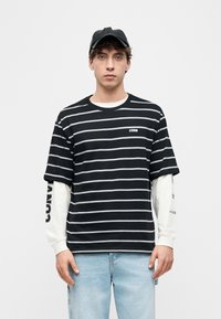 Νέος άνδρας που φορά μαύρο και γκρι κοντομάνικο ριγέ πουκάμισο πάνω από λευκό μακρυμάνικο, ανοιχτόχρωμο τζιν και μαύρο καπέλο, όρθιος μπροστά σε λευκό φόντο.