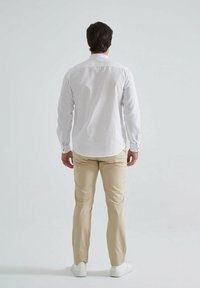 Chemise blanche à manches longues, associée à un pantalon beige et des chaussures blanches, vue de dos sur fond neutre.