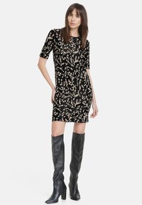 Schwarzes Kleid mit beigem abstraktem Muster, kurzen Ärmeln, figurbetonter Silhouette und knielanger Länge. Getragen mit hohen schwarzen Lederstiefeln.