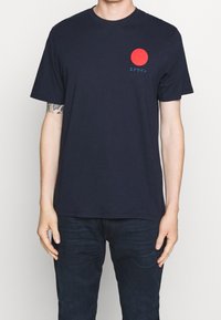 Námořnické bavlněné tričko s kulatou červenou grafikou a japonským textem na levé straně. Krátké rukávy a klasický střih.