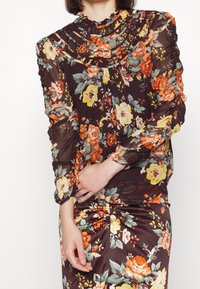 Blouse fleurie en tissu transparent marron avec des motifs orange, jaune et vert, dotée d'un col à volants et de manches longues froncées.