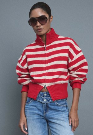 Femme portant de grandes lunettes de soleil foncées, un pull zippé rayé rouge et blanc et un jean bleu taille haute, debout devant un fond gris.