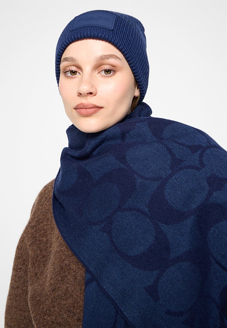 Knitted navy beanie with a logo patch, paired with a matching scarf featuring a tonal circular pattern, draped over a brown sweater.