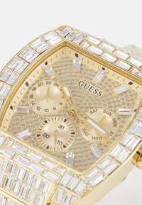 Goldfarbener Uhr mit Kristallverzierungen, rechteckigem Gehäuse, strukturiertem Zifferblatt und Unterziffernblättern für Datum und Tag. Besitzt elegante Stundenmarkierungen.