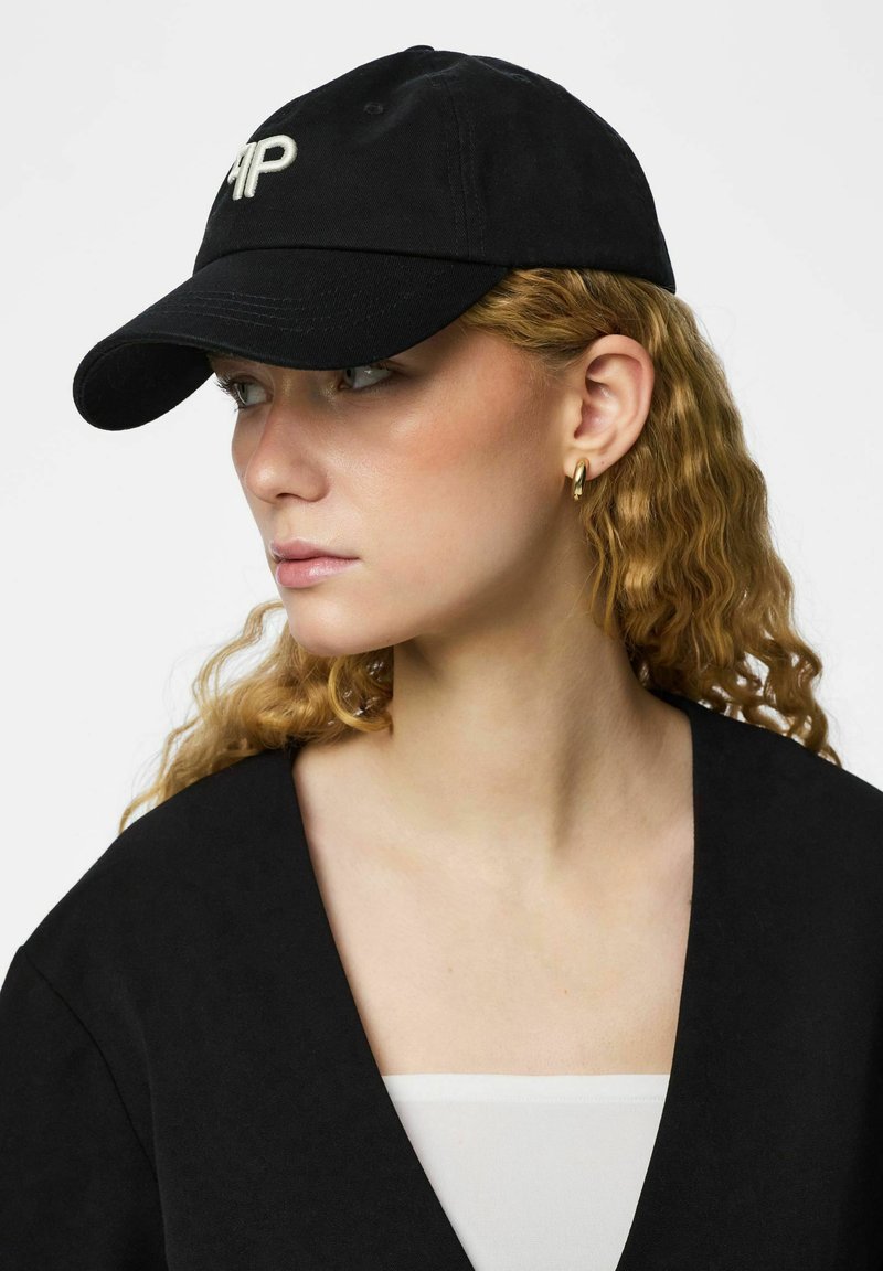 Femme aux cheveux blonds bouclés portant une casquette noire avec un logo blanc, de petites boucles d'oreilles en or, un haut noir et un sous-vêtement blanc, regardant vers la gauche.