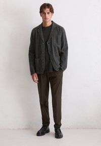 Grijze wollen jas met een kraag met inkeping, voorzak en knoopsluiting, gecombineerd met een donker overhemd en olijfgroene taps toelopende broek. Zwarte schoenen.