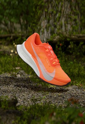 Világos narancssárga futócipő fehér talppal és ezüst Nike logóval, kavicsos ösvény felett lebegve, zöld lombozattal a háttérben.