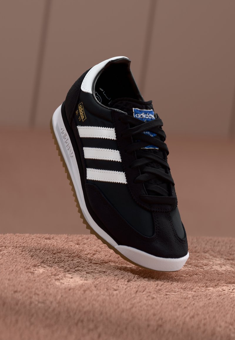 Zwarte sportschoen met witte drie-strepen ontwerp, gemaakt van leer en suede, rubberen zool met een gestructureerd grippatroon en blauwe logo-accent.