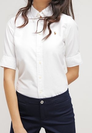 Mujer vistiendo una camisa blanca de manga corta con botones y un pequeño logo bordado, junto con pantalones de color azul marino oscuro.