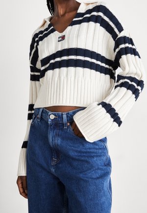 Person iført en hvid og marineblå stribet ribstrikket sweater og højtaljede blå jeans, med hånden i lommen, mod en ensfarvet baggrund.