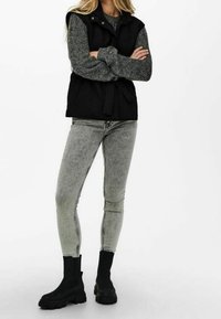 Černá bezrukávová vesta s páskem v pase, kombinovaná se šedým texturovaným svetr a šedými skinny džíny. Černé kotníkové boty outfit doplňují.
