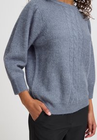 Pull en tricot gris clair avec texture côtelée et motif en cable. Doté de manches trois-quarts et d'une coupe décontractée. Associé à un pantalon noir.