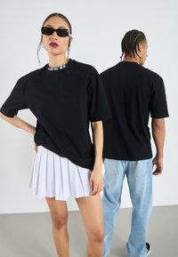Černé oversize tričko s logem „TOKYO“, doplněné bílou plisovanou sukní. Modelka nosí elegantní černé sluneční brýle. Pozadí je neutrální.