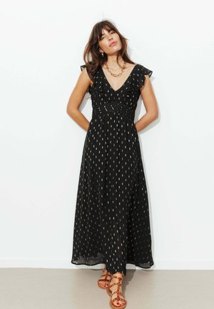 Femme portant une robe maxi noire sans manches avec des motifs rectangulaires dorés et des sandales marron cloutées, debout contre un mur blanc.