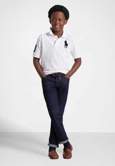 Balta polo krekls ar tumši zilu logotipu, tumšu džinsu bikses ar uzlocītiem aprocēm, brūnas loafers apavi. Modelis stāv ar rokām kabatās, pret gaišu fonu.