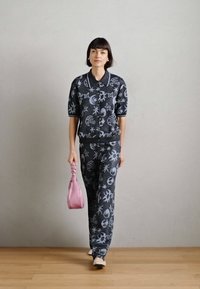 Δύο κομμάτια ρούχων σε σκουρόχρωμο ύφασμα με μωβ γραφικά σχέδια που απεικονίζουν αστέρια και φεγγάρια. Το μοντέλο κρατά μια ροζ, υφασματική τσάντα. Ουδέτερο φόντο.