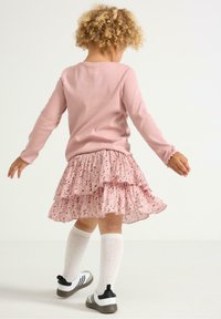 Top a maniche lunghe rosa chiaro abbinato a una gonna a strati con volant e motivo floreale. Il bambino indossa calze bianche fino al ginocchio e scarpe sportive.