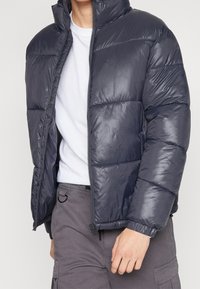 Donkergrijze donsjas van glanzend materiaal, met hoge kraag, ritssluiting en horizontaal doorgestikt patroon. Gecombineerd met een wit overhemd en grijze broek.