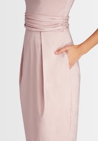 Robe rose pâle avec une taille froncée, une poche latérale et une texture lisse, dotée d'un corsage ajusté et d'une jupe évasée.