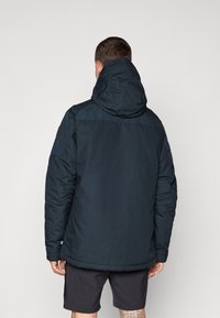 Dark blue insulated jacket with a hood, featuring a smooth texture and a logo patch on the upper arm, and worn over black shorts.